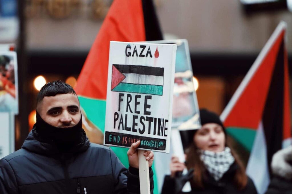 Gambar ini menampilkan dua orang yang memakai kacamata dan memegang bendera Palestina. Mereka berada dalam rally atau protes untuk menentang Israel. Ada juga bendera Palest